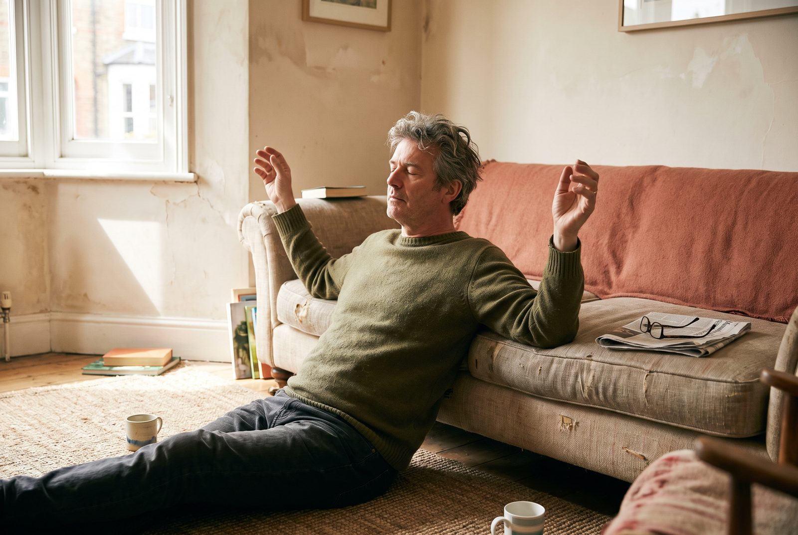 A man in his fifties sits on the floor against a sofa, eyes closed, slowly raising his arms in morning light.